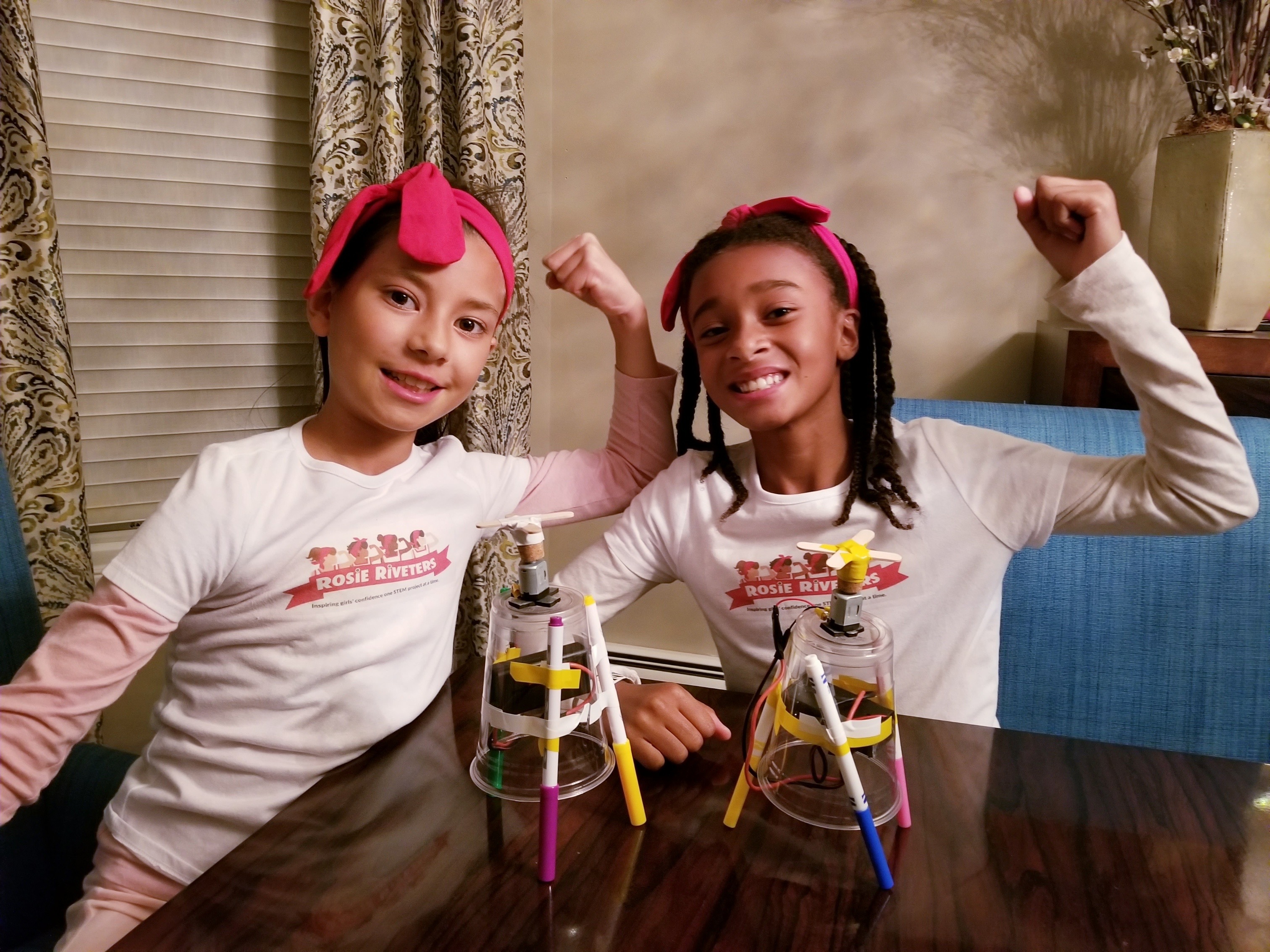 Two girls wearing Rosie the Riveter style bandanas flexing their arms in a Rosie the Riveter post with the two drawing art Robots they built.